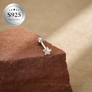 Peut inclure: Boucle d'oreille argentée avec un charm en forme d'étoile. La boucle d'oreille est en argent sterling S925 et possède une petite boule au sommet. L'image affiche la boucle d'oreille sur une surface marron texturée.