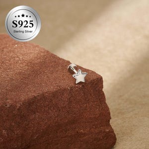 Puede incluir: Un pequeño pendiente plateado en forma de estrella con parte trasera plana y poste. El pendiente está hecho de plata de ley S925. El pendiente se muestra sobre una superficie rugosa de color marrón rojizo. Un logotipo circular con el texto "S925 Sterling Silver" está en la esquina superior izquierda.