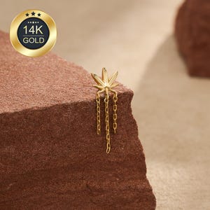 May include: A 14K gold earring featuring a stylized leaf design at the top with three delicate dangling chains extending downward. The piece is crafted from polished yellow gold and displays a detailed, symmetrical leaf shape with pointed edges.