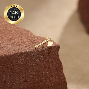 May include: A small 14K gold stud earring shaped like a double-bitted axe. The earring features a polished gold finish with a detailed handle and two symmetrical blade edges. A circular badge in the upper corner displays the text 14K GOLD.