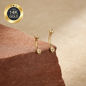 May include: Two curved, gold-colored belly button rings with a small, clear gem at each end. The rings are displayed on a textured, reddish-brown surface. A gold emblem with the text "14K GOLD" is in the upper left corner.
