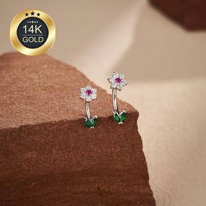 May include: A pair of flower-shaped earrings. Each earring features a white flower with a red center stone, a curved silver stem, and two green leaf-shaped accents. The earrings are set against a brown background, with a gold 14K gold seal in the upper left corner.