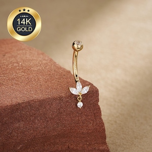 May include: A gold-colored belly button ring with a curved barbell design. The ring features a small, clear gem at the top and a cluster of three white, petal-shaped gems with a dangling round gem at the bottom. The image includes a gold-colored emblem that reads "14K GOLD".