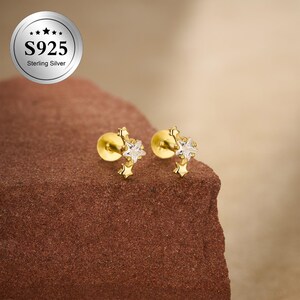 May include: A pair of gold-colored stud earrings. Each earring features a small, round base with a cluster of three stars. The central star is clear, while the others are gold. The earrings are on a brown surface. The image also includes a silver circle with the text "S925 Sterling Silver."