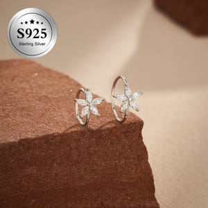 May include: A pair of silver earrings, each with a flower design. The flowers are made of clear gemstones set in a silver hoop. The earrings are displayed on a textured, brown surface. The image includes a circular logo that reads "S925 Sterling Silver."