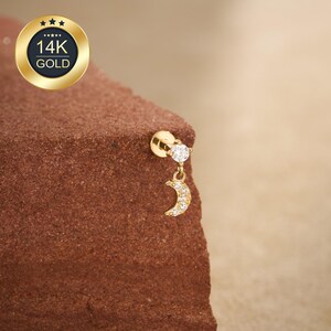 May include: A gold earring with a small, sparkling diamond and a dangling crescent moon shape, also adorned with small, clear stones. The earring is set against a textured, reddish-brown surface. A gold emblem with the text "14K GOLD" is in the upper left corner.