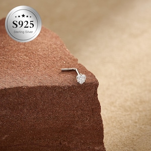 May include: A sterling silver nose stud with a heart-shaped cubic zirconia stone. The jewelry is set on a brown, textured surface. The image also includes a silver circle with the text "S925 Sterling Silver".