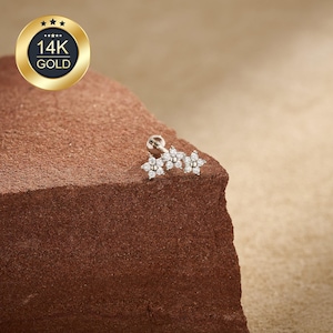 May include: A small, silver-colored earring with three flower-shaped designs, each adorned with small, clear gemstones. The earring is set against a textured, brown surface. A gold-colored emblem with the text "14K GOLD" is in the upper left corner.