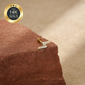 May include: A gold-colored, lightning bolt-shaped earring with small, clear gemstones. The earring is set on a post. A gold-colored emblem with the text "14K GOLD" is in the upper left corner.