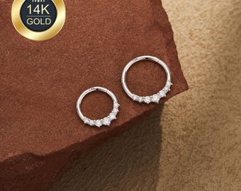 May include: Two silver-colored hoop earrings with a row of small, clear gemstones. The earrings are displayed on a textured, reddish-brown surface. A gold-colored emblem with the text "14K GOLD" is in the upper left corner.