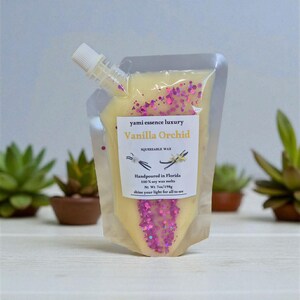Może przedstawiać: Przezroczysty, ściśliwy woreczek z woskiem z białą dyszą, wypełniony żółtym woskiem i różowym brokatem. Etykieta głosi "Vanilla Orchid" i "Handpoured in Florida". Waga netto wynosi 198g.
