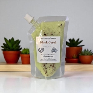 May include: A clear, squeezable wax pouch filled with light green wax, speckled with brown particles. The label reads "Black Coral" and "Handpoured in Florida." The pouch has a white spout and is displayed in front of potted succulents.