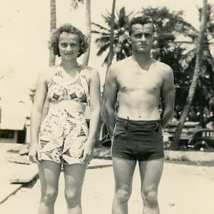May include: A vintage black and white photograph showing two individuals standing side by side. One wears a two-piece swimsuit with a patterned halter top and matching high-waisted shorts. The other wears dark, solid-colored swim trunks.