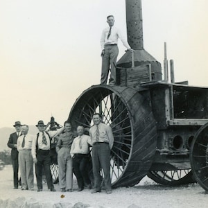 May include: A vintage black and white photograph of a group of men posing with a large, antique tractor. The tractor has large spoked wheels and a tall smokestack. The men are dressed in shirts and ties, and some wear hats.