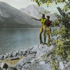 May include: A vintage colorized photograph showing two individuals standing on a rocky lakeshore. One wears a lime green short-sleeved shirt and tan trousers, while the other wears a dark red shirt and tan trousers, pointing toward mountains.