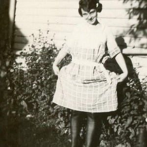 May include: A vintage black and white photograph of a person wearing a plaid dress with a white belt and dark boots. The person is standing in front of a bush and a wooden wall.