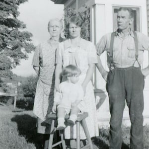 May include: A black and white photo of four people. Two women and a man are standing, while a young child sits on a stool. The women are wearing dresses and the man is wearing a collared shirt and overalls. The child is wearing a dress and has short hair.