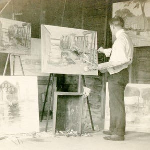 Peut inclure: Photographie en noir et blanc d'un artiste dans son atelier. L'artiste peint un paysage sur un chevalet. Plusieurs autres peintures sont visibles en arrière-plan, créant une ambiance artistique et créative.