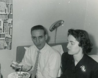 May include: Black and white photograph of a man and a woman. The man is holding a plate of food and a sandwich. The woman is wearing a dark jacket with a brooch. The photo appears to be taken indoors.
