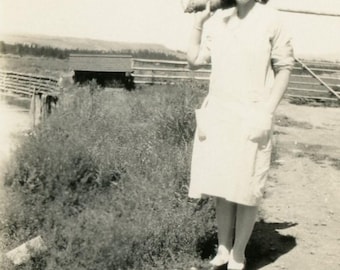 May include: A vintage black and white photograph of a person wearing a light-colored dress with pockets, standing outdoors. The person is smiling and carrying an object over their shoulder. A rural landscape with a fence and building is in the background.