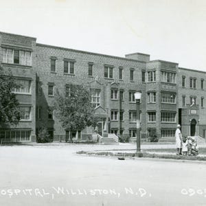 Puede incluir: Fotografía en blanco y negro de un gran edificio de ladrillo, probablemente un hospital, con múltiples ventanas y una entrada central. La imagen incluye una calle, una farola y personas cerca de la entrada. El texto en la parte inferior dice "HOSPITAL, WILLISTON, N.D."
