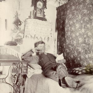 May include: A vintage sepia-toned photograph showing a person reclining on a bed, holding a pipe and a cat. Beside the bed is a sewing machine table with a lace cover, a wall clock, and a floral patterned curtain hanging in the background.