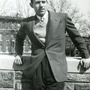 May include: A black and white photograph of a person wearing a dark blazer over a sweater and collared shirt. The person is leaning on a stone wall, with a building and trees in the background.