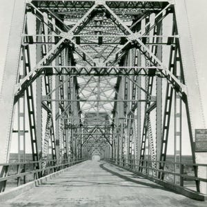 Puede incluir: Fotografía en blanco y negro de un puente de armadura de acero. El puente tiene una compleja red de vigas y soportes metálicos, con una carretera que atraviesa el centro. El puente se extiende a lo largo de una distancia, con la carretera desapareciendo en la distancia.