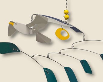 May include: A metal mobile featuring a seagull with white, gray, and blue wings, a yellow beak, and a yellow ring. Below, teal leaf-shaped elements hang from thin wires. Two yellow spheres and a hook complete the design.