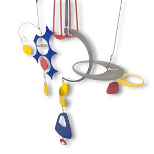 May include: A colorful, abstract mobile sculpture with geometric shapes. The mobile features red, blue, yellow, and gray elements, including circles, teardrops, and curved forms, suspended by wires. The artwork is designed to hang and rotate.