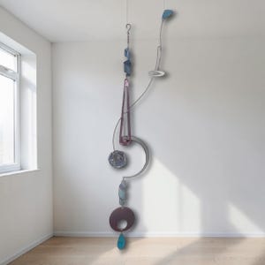 May include: Abstract mobile sculpture with a mix of shapes and colors. Features a series of geometric forms, including a crescent, a teardrop, and a triangle, in shades of purple, blue, and silver. The mobile hangs from a thin wire.