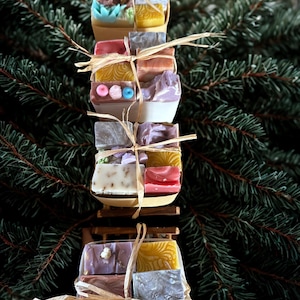 Könnte beinhalten: Ein Stapel handgemachter Seifen in verschiedenen Farben, zusammengebunden mit Bast. Die Seifen sind auf kleinen Holzplattformen angeordnet, mit einem Hintergrund aus grünen Tannennadeln. Die Seifen haben dekorative Elemente und unterschiedliche Texturen.