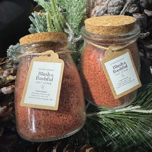 May include: Two glass jars of pink salt scrub with cork lids and tags reading "Blush & Bashful Salt Scrub." The jars are displayed amongst pine needles and decorative foliage, creating a natural aesthetic.