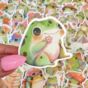Puede incluir: Una pegatina de rana verde con manchas marrones y grandes ojos negros que sostiene una flor rosa. La pegatina tiene un borde blanco y está rodeada de otras pegatinas de ranas con varios accesorios.