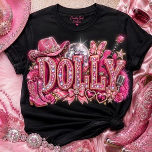 May include: A black t-shirt featuring the word DOLLY in large, pink, rhinestone-encrusted letters. The graphic includes a pink cowboy hat, a disco ball, a guitar, a microphone, butterflies, roses, and a bow. The brand name Country Girl Couture is printed at the neckline.