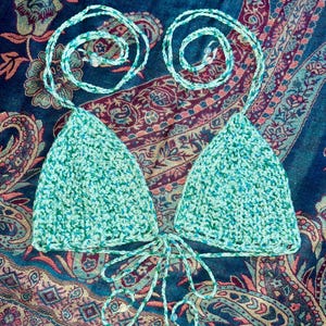 May include: A crochet bikini top featuring two triangular cups in a variegated mint green and teal yarn. The garment includes thin braided neck and back ties, each finished with small clear beads at the ends, creating a textured handmade appearance.