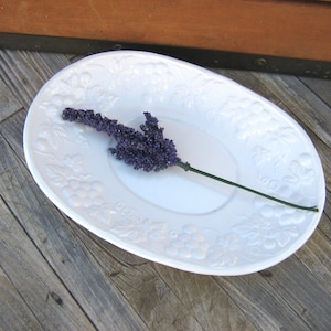 May include: White oval serving dish with a raised floral design. A sprig of purple flowers is resting on the dish.