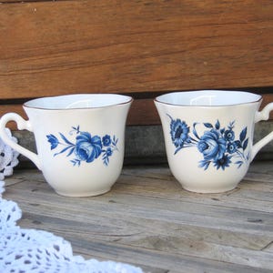 May include: Two white teacups with blue floral designs. The cups have a scalloped rim and a handle.