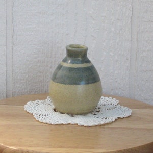 May include: A small, hand-thrown ceramic vase with a light brown body and a blue and white stripe design. The vase is sitting on a wooden table with a white doily.