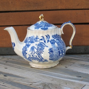 May include: White and blue ceramic teapot with a lid, featuring a detailed blue willow pattern. The teapot has a curved spout, a handle with gold accents, and a gold finial on the lid. It sits on a wooden surface.