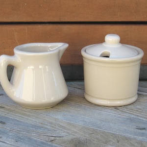 May include: A set of two off-white ceramic kitchenware items. A small creamer pitcher with a handle and a lidded sugar bowl.
