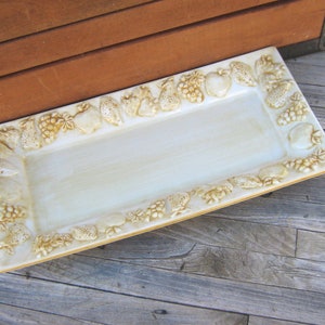 May include: A rectangular ceramic serving tray with a raised, embossed design of fruit and grapes around the edge. The tray is white with a light brown glaze.