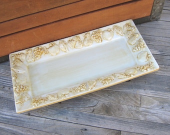May include: A rectangular ceramic serving tray with a raised, embossed design of fruit and grapes around the edge. The tray is white with a light brown glaze.