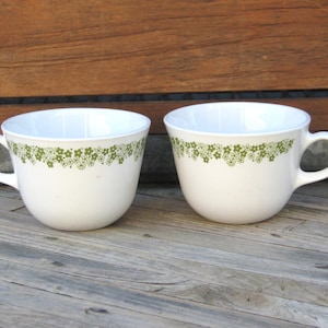 May include: Two white ceramic mugs with a green floral pattern around the rim.