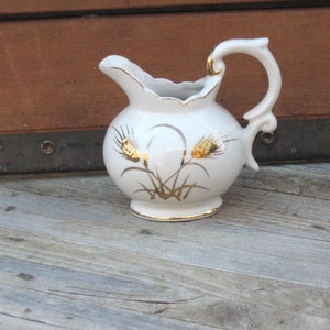 May include: White ceramic pitcher with a gold wheat design. The pitcher has a handle and a spout.