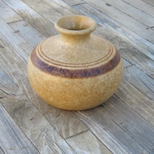 May include: A small, round, tan ceramic vase with brown stripes. The vase is sitting on a wooden surface.