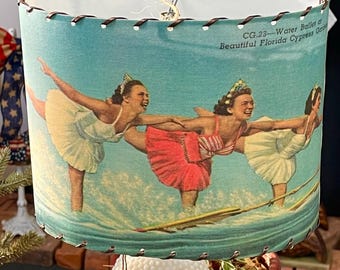 May include: A vintage table lamp with a blue lampshade featuring a water ballet scene. The lamp base is white with a textured, hobnail design. The shade has the text "CG 23 - Water Ballet of Beautiful Florida Cypress Gardens."
