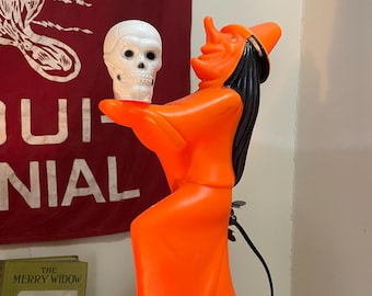 May include: A vintage Halloween decoration featuring an orange witch holding a white skull. The witch has black hair and a pointed hat. The decoration is placed on top of old books. A red banner with white text is in the background.
