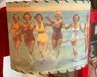 May include: A vintage lampshade featuring a retro illustration of women in swimsuits running in the ocean. The lampshade has a stitched edge and is mounted on a dark wooden lamp base. The text "BEAUTIES ON THE BEACH" is visible.