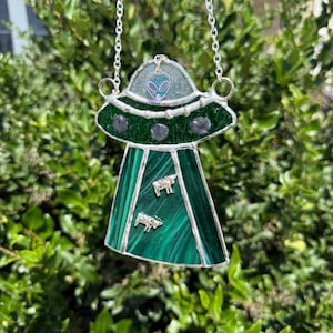Op de afbeelding: Een gebrandschilderd glazen UFO-zonnevanger met een zilveren ketting. Het groene en heldere glazen ontwerp heeft een alien, grijze stenen en twee koeienbedels. De zonnevanger wordt omhoog gehouden tegen een achtergrond van groen gebladerte.
